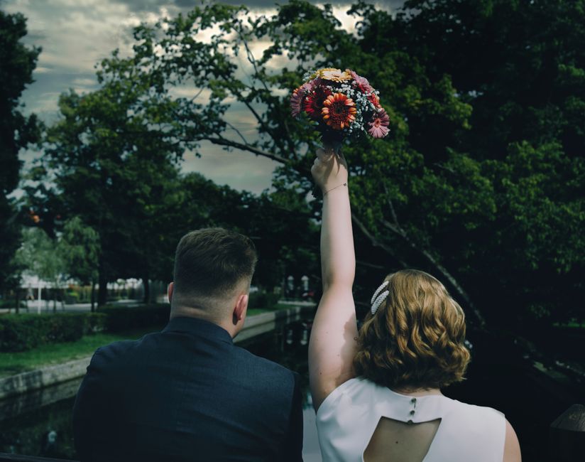 A woman with curly, shoulder-length reddish-brown hair, wearing a white sleeveless dress with a keyhole back detail, is holding a vibrant bouquet of fresh flowers high in the air during an outdoor wed