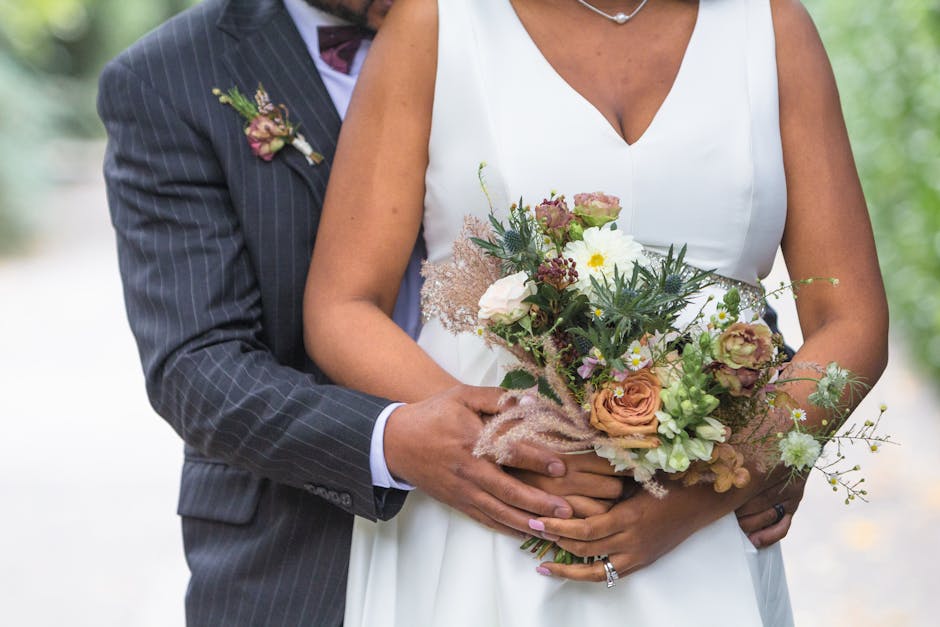 A bride and groom holding a bouquet of fresh flowers in soft peach, white, and green hues, including roses, daisies, and greenery, arranged in a natural, loosely styled floral gift. The bouquet is wra