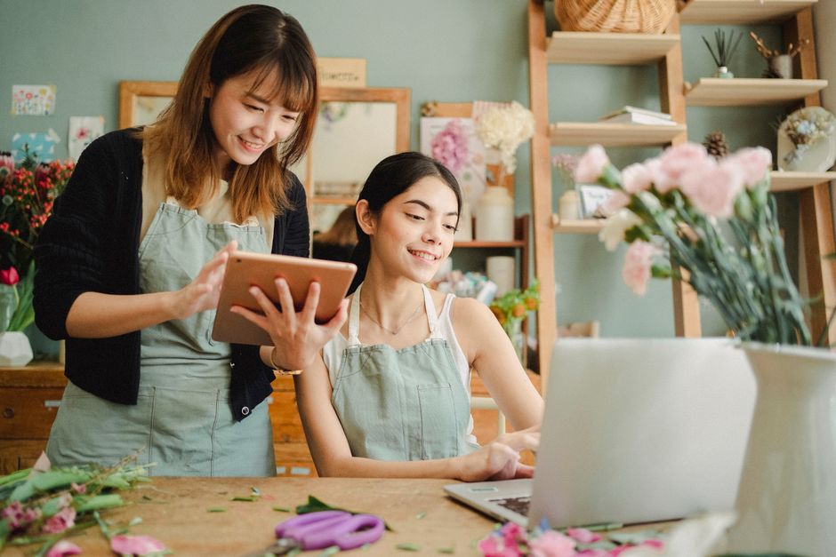 Two women working in a flower shop, one standing and holding a tablet and the other sitting at a wooden table with a laptop, arranging and examining a bouquet of fresh pink and white roses, carnations