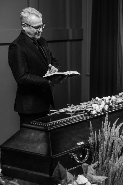 A black-and-white photograph showing a man with gray hair and glasses dressed in a dark suit, standing beside a closed casket decorated with white and pale pink roses. The man is holding a book or not