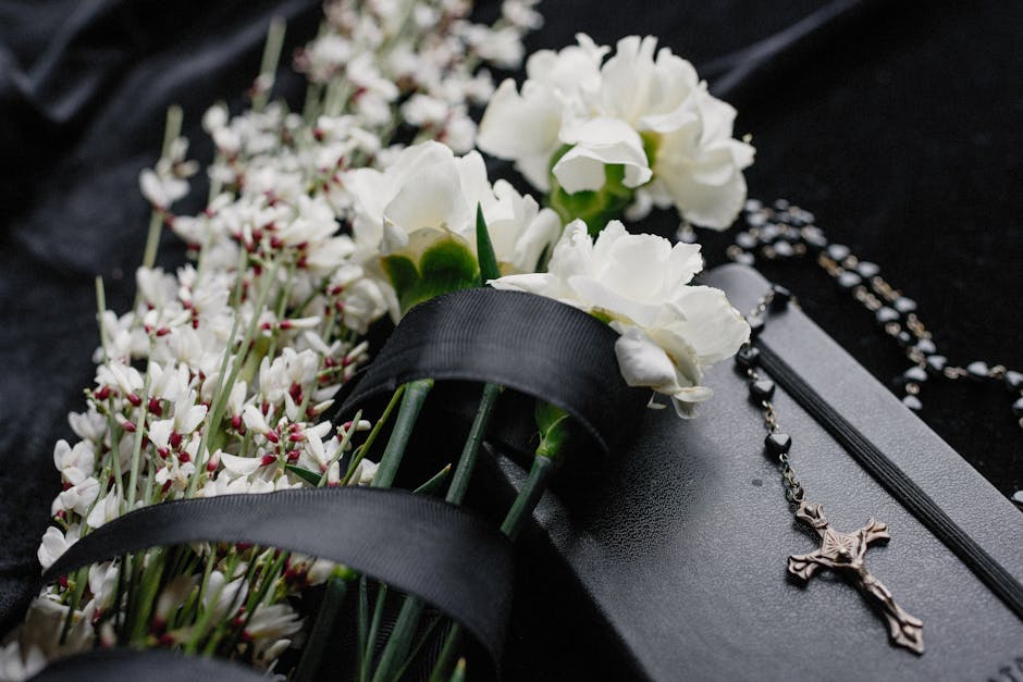 Close-up of white carnations and small white flowers with pink accents, arranged on a black surface, partially wrapped in black satin ribbon. Beside the flowers, there is a black prayer book and a sil