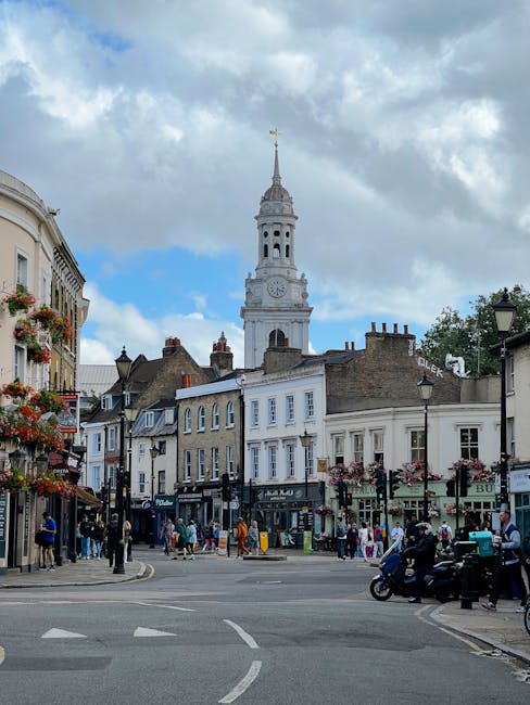 Photograph of a bustling city street with historic-style buildings and storefronts, adorned with flower baskets and hanging flower displays. In the background, a tall, white clock tower with a spire a